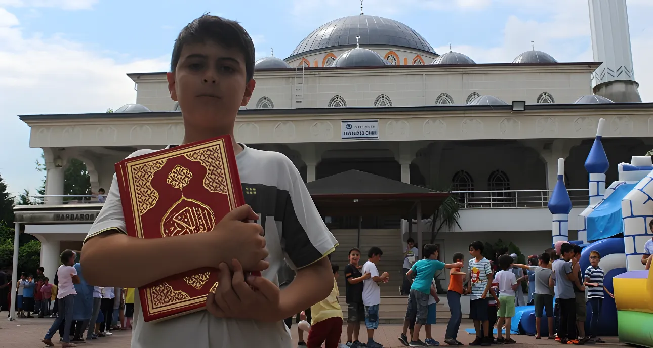 A young boy holding a Quran in front of a mosque.